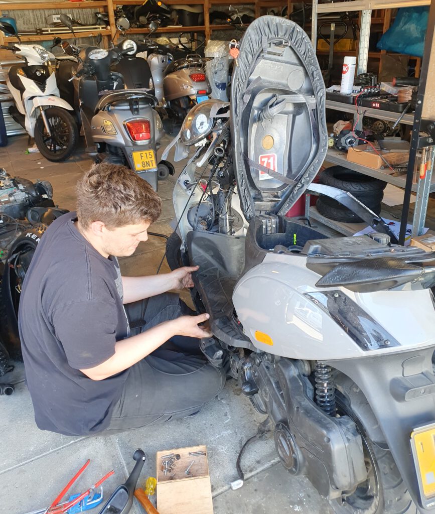 Monteur Roland van Bromfix voert een reparatie uit aan een grijze Piaggio Zip in de werkplaats in Berkel en Rodenrijs. Op de foto is te zien hoe de gaskabel wordt vervangen en de kappen zijn gedemonteerd voor technisch onderhoud aan deze Piaggio scooter.