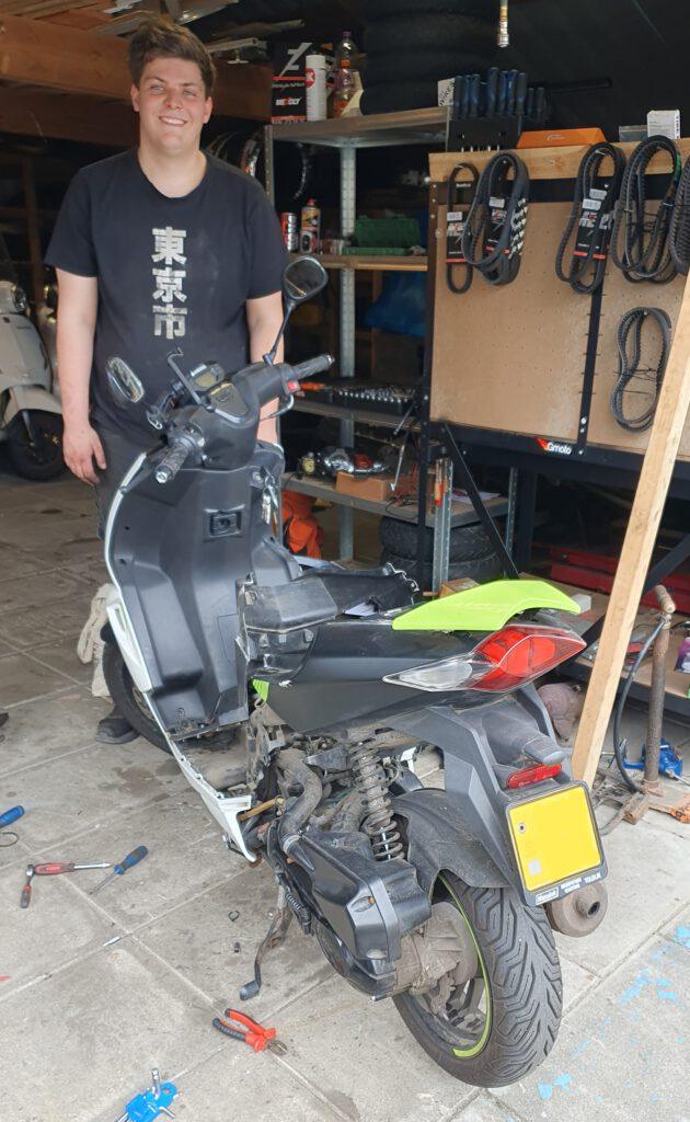 Roland, monteur van Bromfix in de werkplaats in Berkel en Rodenrijs tijdens de reparatie van een Peugeot Speedfight 4 scooter met een gebroken remkabel. Op de achtergrond is een georganiseerd onderdelenbord met v-snaren zichtbaar.
